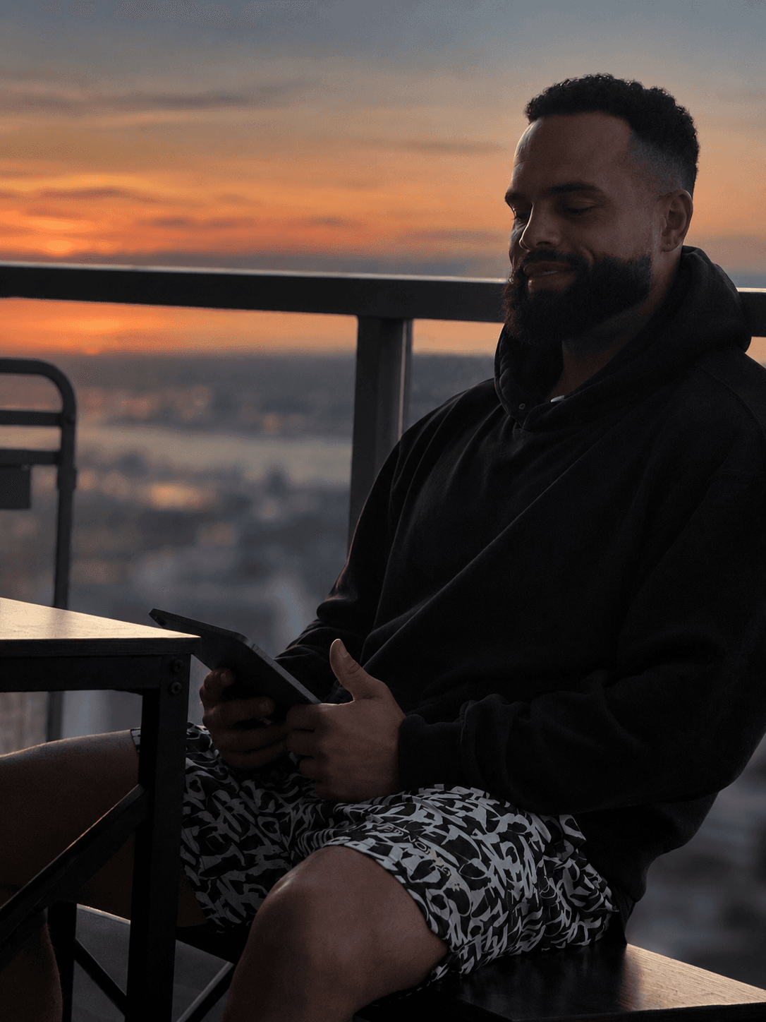 Andrew Paul in a black hoodie holding his tablet on a balcony at sunset